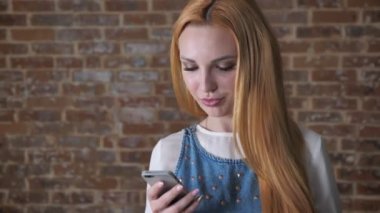 Genç çekici sarışın kız gülümsüyor, tuğla arka plan, iletişim kavramı smartphone, mesaj okuyor