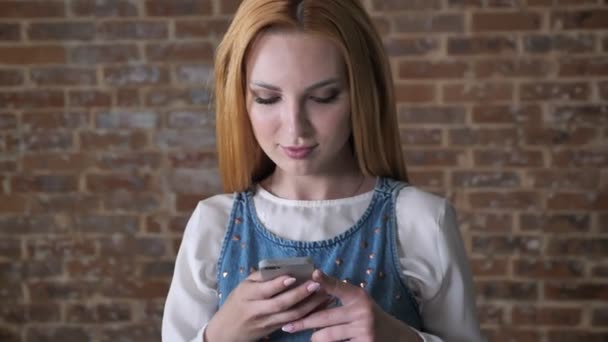 Joven chica rubia seductora está escribiendo mensaje en el teléfono inteligente, fondo de ladrillo, concepto de comunicación — Vídeo de stock