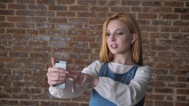 Joven chica rubia dulce tiene una videollamada en el teléfono inteligente, fondo de ladrillo, concepto de comunicación — Vídeos de Stock