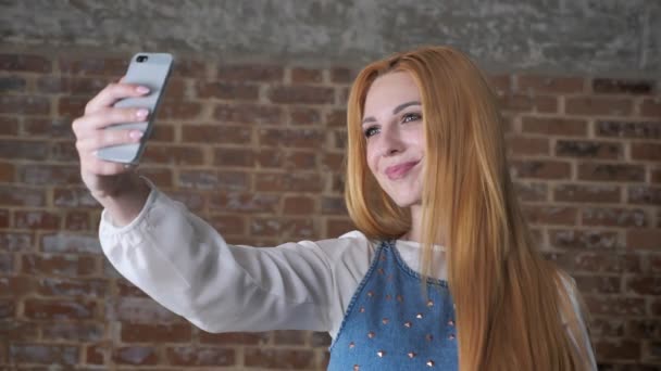 Young sweet blonde girl is making selfie on smartphone, smiling, brick background, communication concept — Stock Video