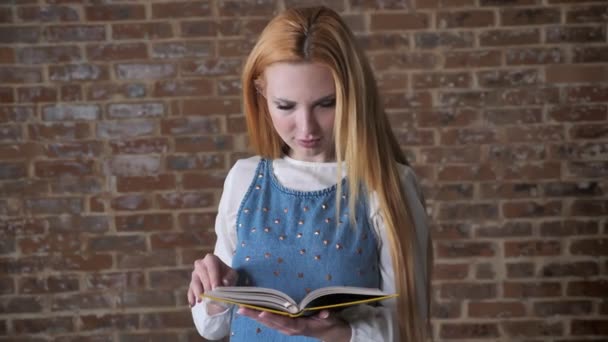 Young alluring blonde girl is reading book, watching at camera, smiling, brick background — Stock Video