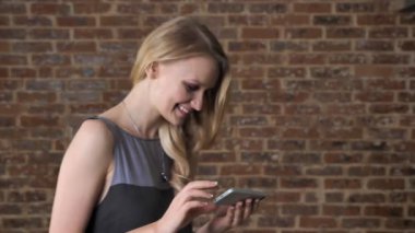 Genç Sevimli sarışın kız gülümsüyor, iletişim kavramı, tuğla arka plan fotoğraf Smartphone, izliyor