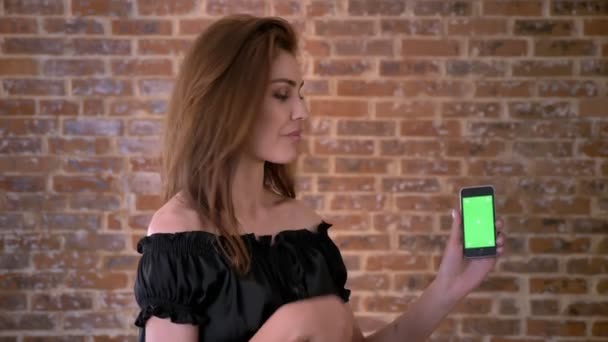 Charmante femme montre l'écran vert du smartphone, comme signe, concept de communication, fond de brique — Video
