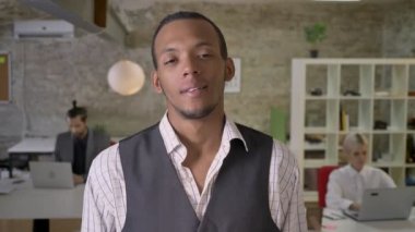 Genç Afrika işadamı duruyor ve ofis, kameraya gülümseyen, izlerken meslektaşları ile dizüstü bilgisayarlar, iş kavramı çalışıyoruz