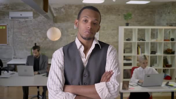 Young african businessman is standing with arms crossed and watching at camera in office, thinking concept, colleagues are working with laptops, work concept — Stock Video