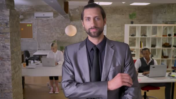 Homme d'affaires avec barbe est debout avec les bras croisés et regarder à la caméra dans le bureau, collègues travaillent avec des ordinateurs portables, concept de travail — Video
