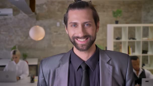 Hombre de negocios guapo con barba está mirando a la cámara en la oficina, sonriendo, los colegas están trabajando con computadoras portátiles, concepto de trabajo — Vídeo de stock