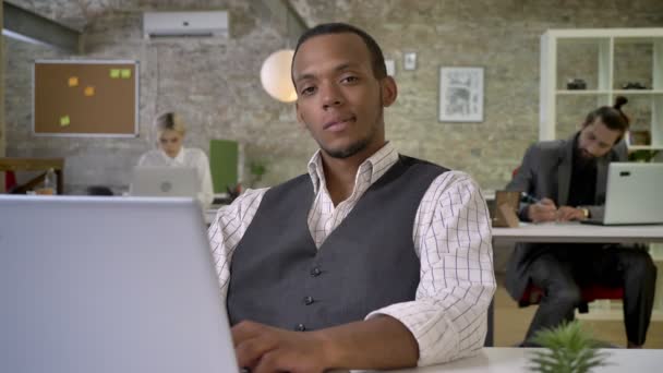 Jovem empresária africana está sentada no laptop no escritório, assistindo à câmera, colegas estão trabalhando em rede com tecnologias, conceito de trabalho, conceito de comunicação — Vídeo de Stock