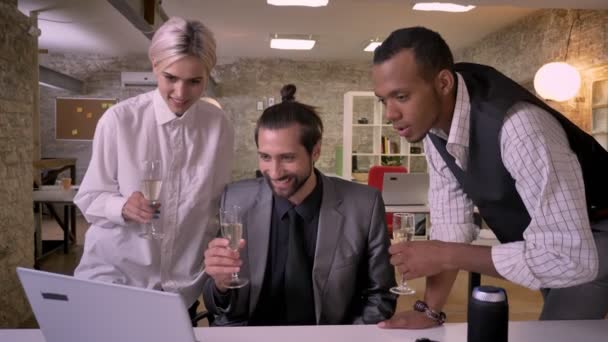 Tres trabajadores multy-étnicos están viendo en la pantalla de la computadora portátil, vasos Tink, beber champán, concepto de fiesta de la empresa, concepto de comunicación — Vídeo de stock