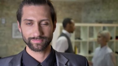 Sakallı yakışıklı emin işadamı kamera ofis, izlerken gülümsüyor, meslektaşları arka planda, iletişim kavramı konuşuyorsun