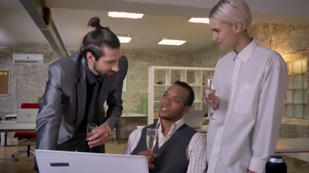 Tres trabajadores multiétnicos cuentan historias, se sientan en la computadora portátil en la oficina, beber champán, concepto de comunicación — Vídeos de Stock
