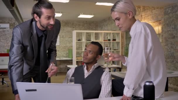 Три багатоетнічні працівники розповідають історії, сидять біля ноутбука в офісі, п'ють шампанське, концепція спілкування, концепція розслаблення — стокове відео