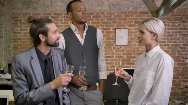 Трое мультиэтнических работников рассказывают истории, шутят, сидят в офисе, пьют шампанское, общаются — стоковое видео