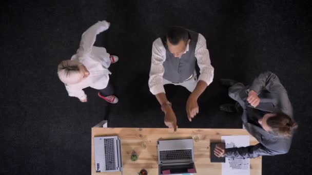 Tres alegres trabajadores multy-étnicos están bailando cerca de la mesa con computadoras portátiles y copas con champán en la oficina, concepto de fiesta de la empresa, top shot — Vídeos de Stock