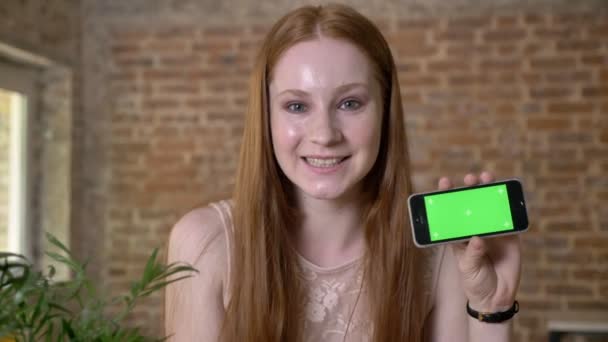 Joven chica pelirroja fresca está mostrando la pantalla verde en su teléfono inteligente, mirando a la cámara, concepto de comunicación, fondo de ladrillo — Vídeo de stock