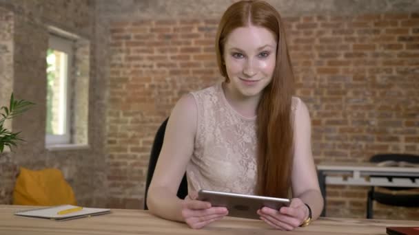 Chica de jengibre linda joven está mostrando la pantalla verde en su tableta, mirando a la cámara, concepto de comunicación, fondo de ladrillo — Vídeos de Stock