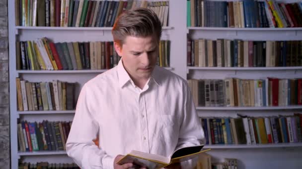 Молодой сконцентрированный мужчина читает книгу в библиотеке, смотрит в камеру — стоковое видео