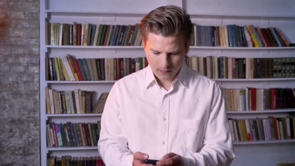 Young man is showing green screen of smartphone in library, watching at camera, communication concept — Stock Video