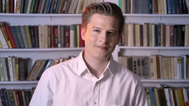 Молодой симпатичный человек смотрит на камеру, библиотека на заднем плане — стоковое видео