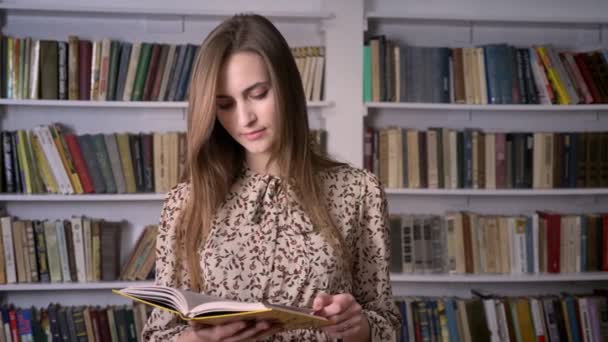Nuori kaunis nainen lukee kirjaa, katselee kameralla, kirjasto taustalla — kuvapankkivideo