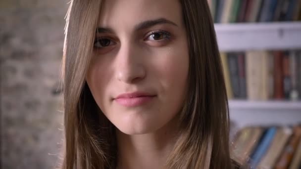 Young serious woman is watching at camera, bookshelf on background — Stock Video