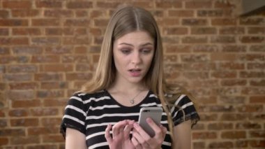 Genç tatlı blonda fotoğraflar smartphone, sürpriz, iletişim kavramı, tuğla arka plan üzerinde izliyor