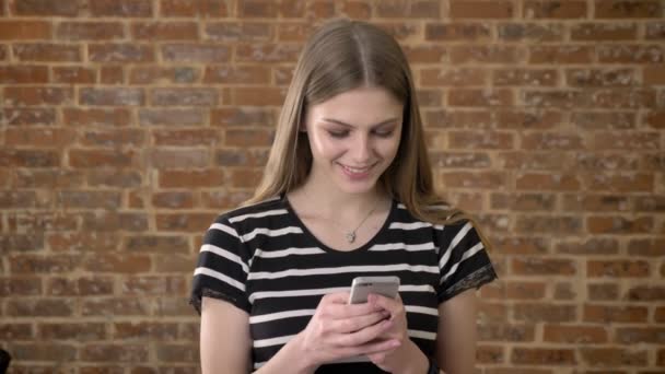 Jeune blonda douce tape message sur smartphone, regarder à la caméra, sourire, concept de communication, fond de brique — Video