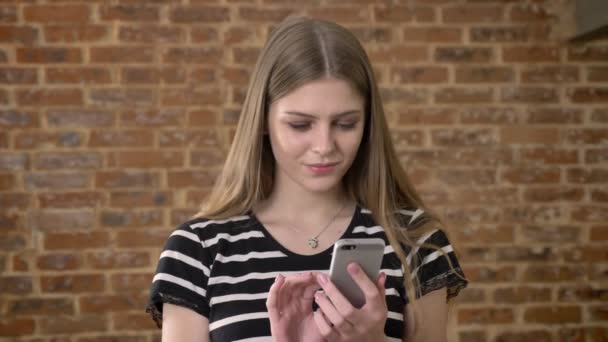 Jeune blonda douce tape message sur smartphone, sourire, concept de communication, fond de brique — Video