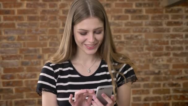 Jeune blonda douce regarde des photos sur smartphone, rire, concept de communication, fond de brique — Video
