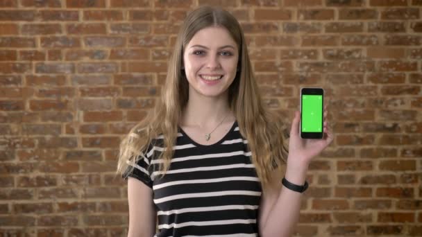 Junge glückliche Blondine zeigt grünen Bildschirm des Smartphones, zeigt darauf, Kommunikationskonzept, Backsteinhintergrund — Stockvideo