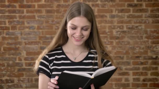 Fiatal gyönyörű blonda az olvasó a könyv, a kamerát, mosolyogva, tégla háttér — Stock videók