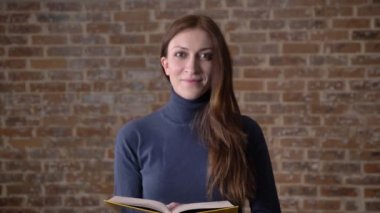 Mutlu kumral kız kameraya, gülümseyen, tuğla arka plan izlerken kitap okuma