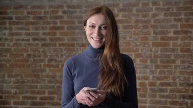 Kumral kız kameraya, gülümseyen, iletişim kavramı, tuğla arka plan izlerken smartphone, ileti yazmakta olduğunu