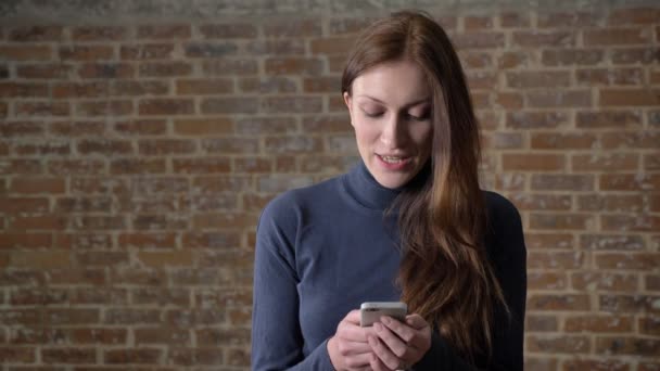 갈색 머리 여자는 입력 메시지 스마트폰, 웃 고, 커뮤니케이션 개념, 벽돌 배경 — 비디오