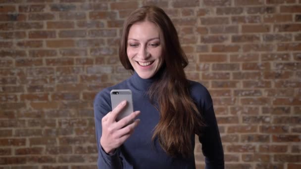 Barna hajú lány videochat smartphone, a kommunikáció fogalma, a tégla háttér beszél — Stock videók