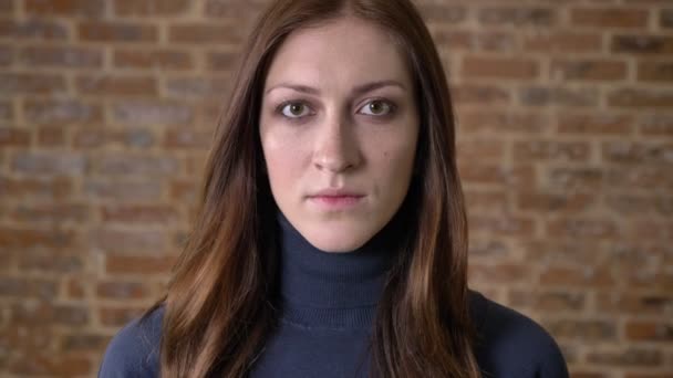 Serious brown hair girl is watching at camera, brick background — Stock Video