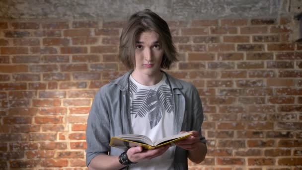 Young long hair man with nose ring is reading book, watching at camera, brick background — Stock Video