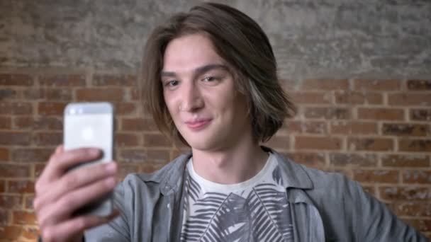 Joven hombre de pelo largo con anillo en la nariz está haciendo vídeo en el teléfono inteligente, saludando, mostrando gesto de paz, concepto de comunicación, fondo de ladrillo — Vídeos de Stock