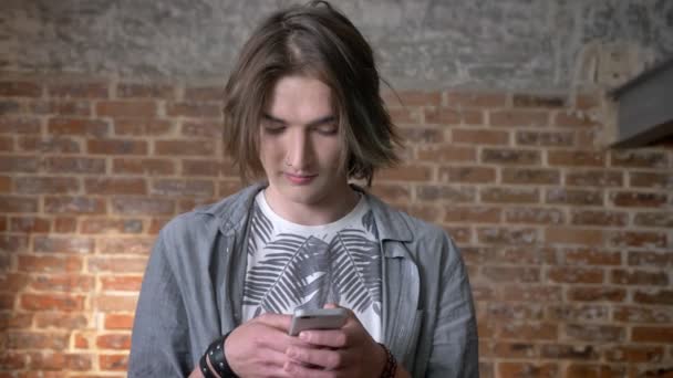 Jonge lange haren man met neusring is bericht aan het typen op smartphone, kijken op camera, baksteen achtergrond — Stockvideo