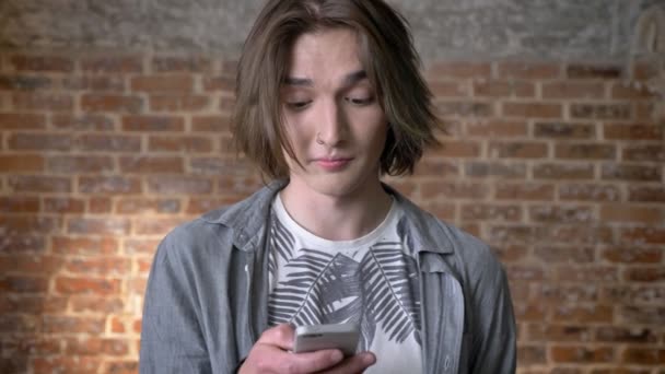 Giovane uomo capelli lunghi con anello naso sta scorrendo le foto su smartphone, guardando la fotocamera, sfondo di mattoni — Video Stock