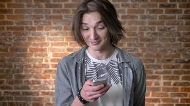 Jonge lange haren man met neusring is typing message op smartphone, glimlachen, communicatieconcept, baksteen achtergrond — Stockvideo