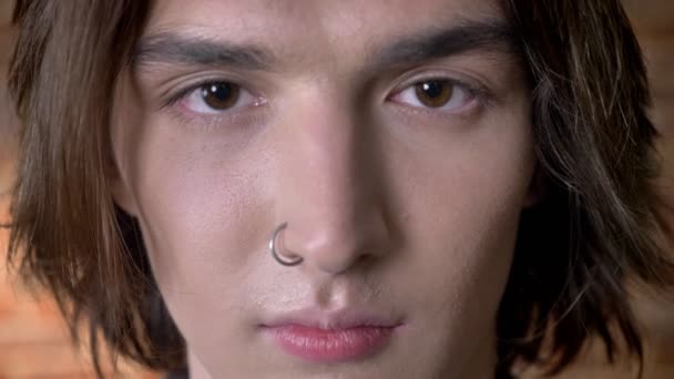 Cara de agradable joven hombre de pelo largo con anillo en la nariz está mirando a la cámara — Vídeo de stock