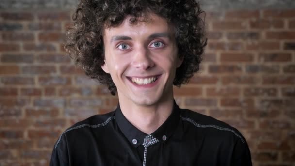 Jovem homem feliz com cabelo encaracolado olhando para a câmera e rindo, fundo da parede de tijolo — Vídeo de Stock