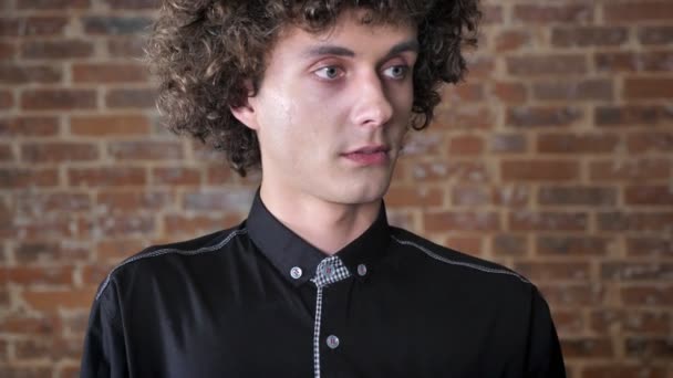 Young surprised man with curly hair raising his eyebrows and looking at camera, brick wall background — Stock Video