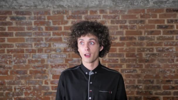 Junger Mann mit lockigem Haar, der mit einer Idee oder Lösung aufwartet, mit erhobenem Zeigefinger und lächelndem, glücklichem Gesichtsausdruck, Backsteinmauer-Hintergrund — Stockvideo