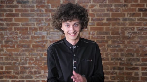 Junger überraschter Mann mit lockigem Haar klatscht und blickt mit erstaunter Miene in die Kamera, Backsteinmauer Hintergrund — Stockvideo