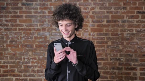 Молодой красивый мужчина с вьющимися волосами, используя свой телефон и стоя возле кирпичной стены — стоковое видео