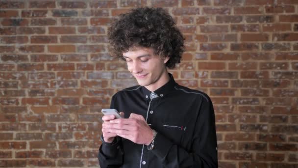 Giovane uomo felice con i capelli ricci chatta al telefono e sorridente, mattone muro di sfondo — Video Stock