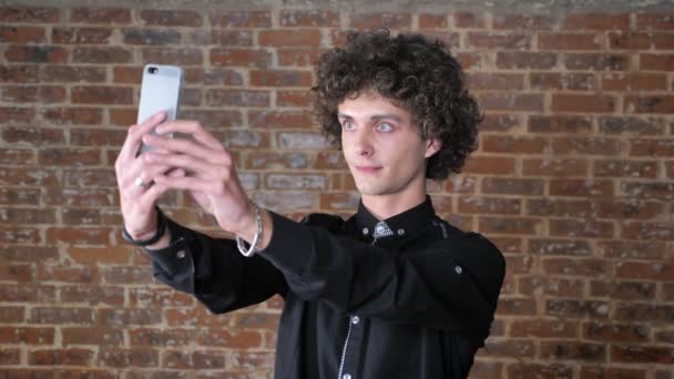 Młody człowiek uśmiechający się z kręconymi włosami biorąc selfie i stojący w pobliżu mur z cegły, wyrażenie zadowolony — Wideo stockowe