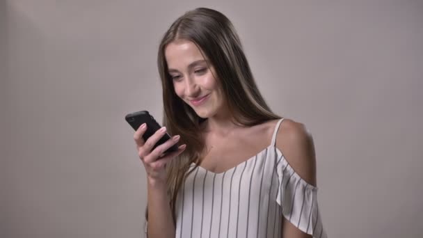 Młoda atrakcyjna dziewczyna nieśmiały pisania wiadomości na smartfonie, oglądając aparatu, uśmiechnięty, komunikacja koncepcja, szare tło — Wideo stockowe